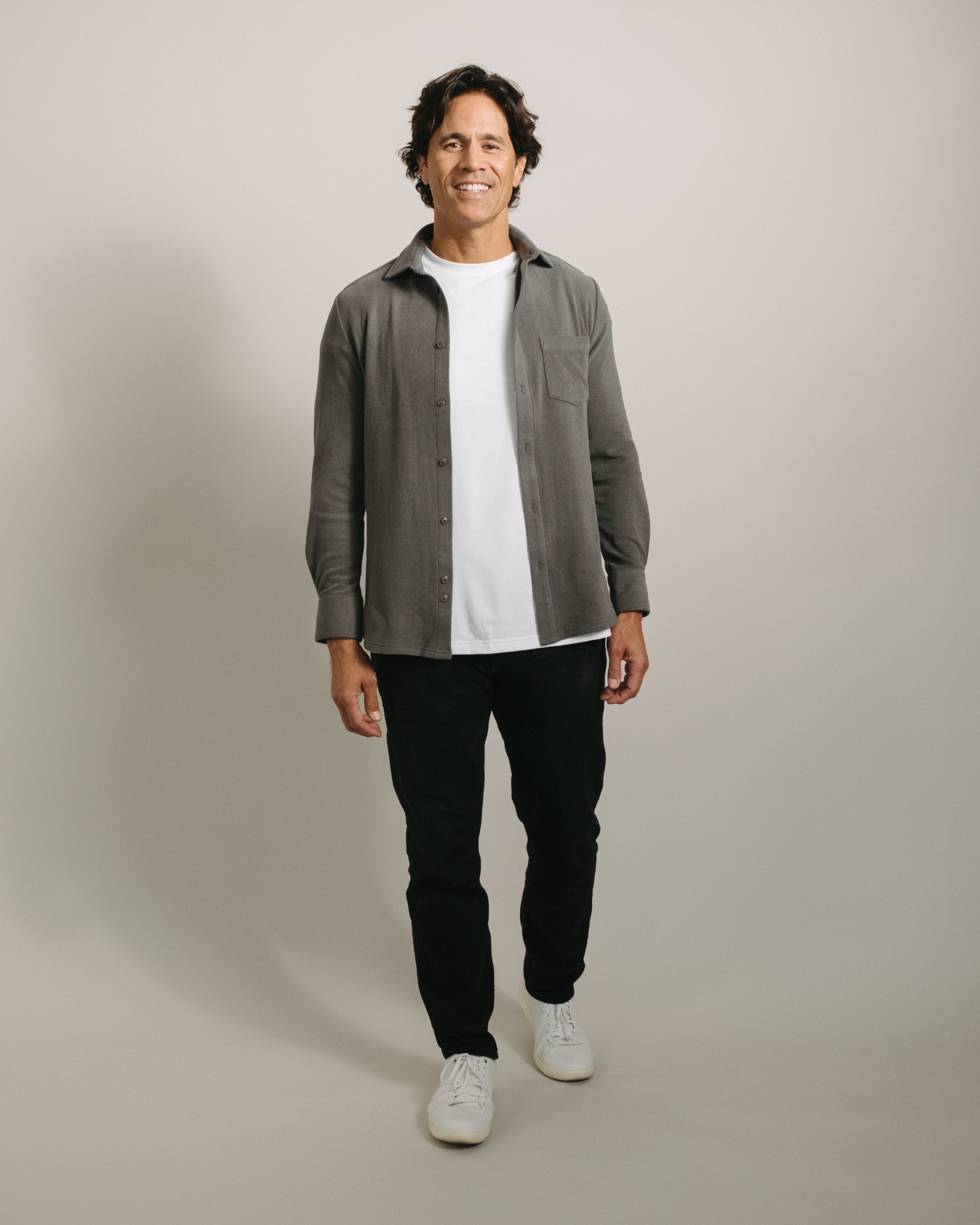 Man wearing a charcoal gray button-up flannel shirt over a white t-shirt, black pants, and white sneakers on a plain background